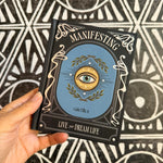 A hand holds a hardcover book titled 'Manifesting' by Gina Elliss, featuring a blue central illustration with an eye and gold laurel wreath, set against a black and white geometric patterned background.