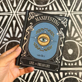 A hand holds a hardcover book titled 'Manifesting' by Gina Elliss, featuring a blue central illustration with an eye and gold laurel wreath, set against a black and white geometric patterned background.