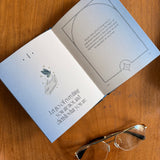 An open book or booklet laid on a wooden table, showing printed text and a small illustration on the pages. A pair of eyeglasses rests near the bottom right corner of the book.