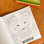 A coloring book page featuring a detailed black and white line art illustration of a Capricorn zodiac sign, styled on a wooden table with colored pencils.