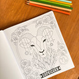 A coloring book page featuring a detailed black and white line art illustration of a Capricorn zodiac sign, styled on a wooden table with colored pencils.