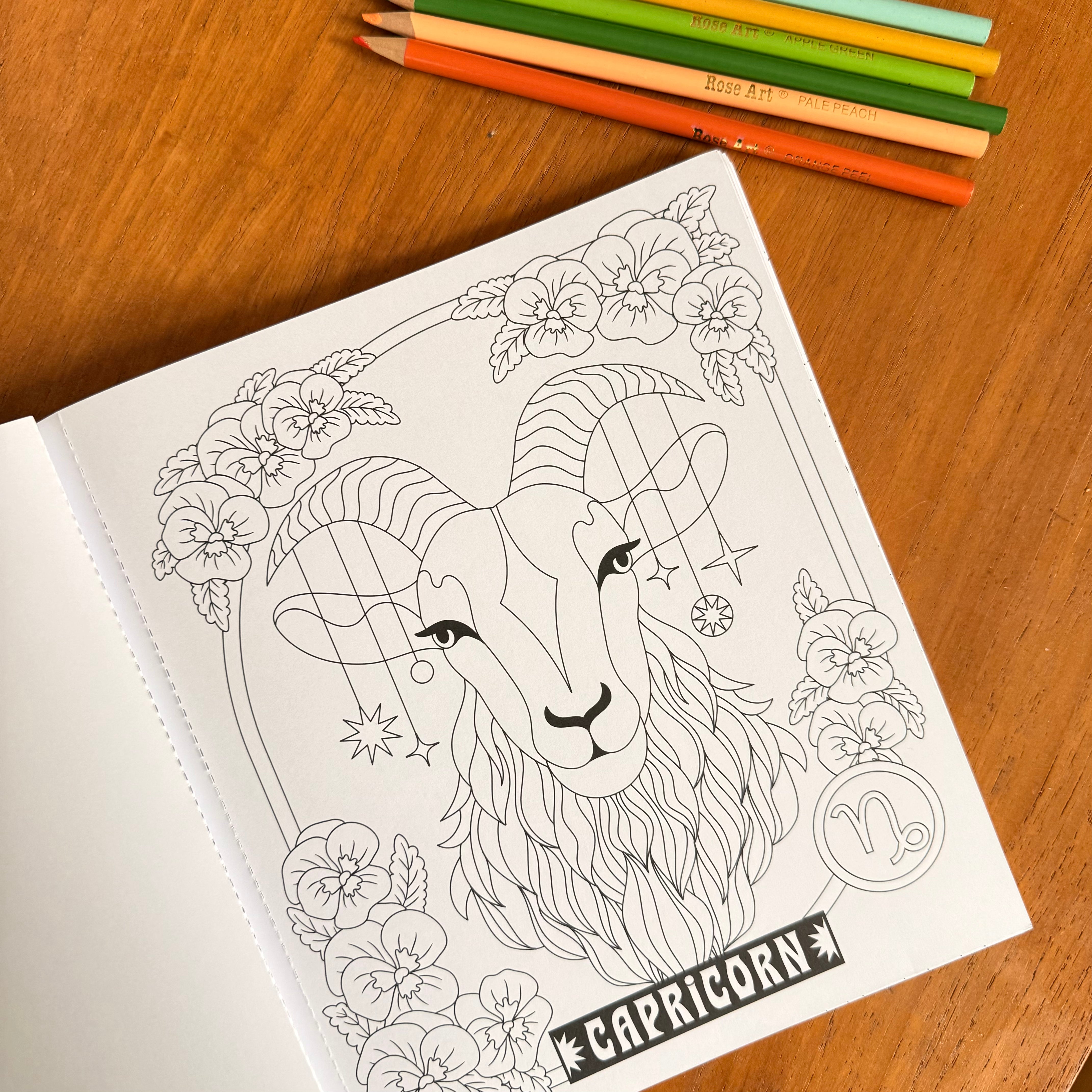 A coloring book page featuring a detailed black and white line art illustration of a Capricorn zodiac sign, styled on a wooden table with colored pencils.