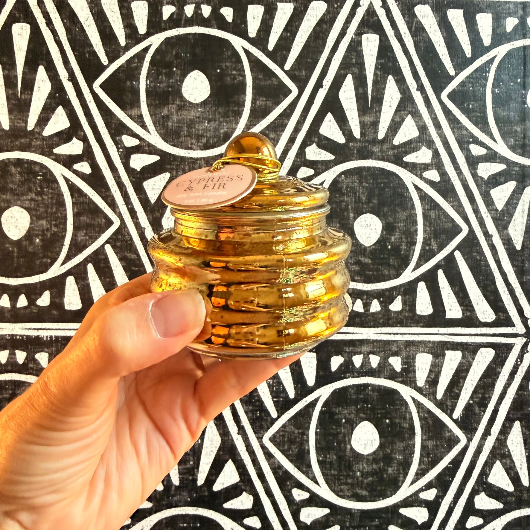 A hand holding a shiny gold vessel with Paddywax Cypress & Fir label against a black and white eye background