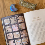 Open book on a wooden surface with a smudge stick and moon-shaped candle