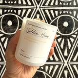 Hand holding a candle labeled 'Golden Hour' against a black and white geometric patterned background