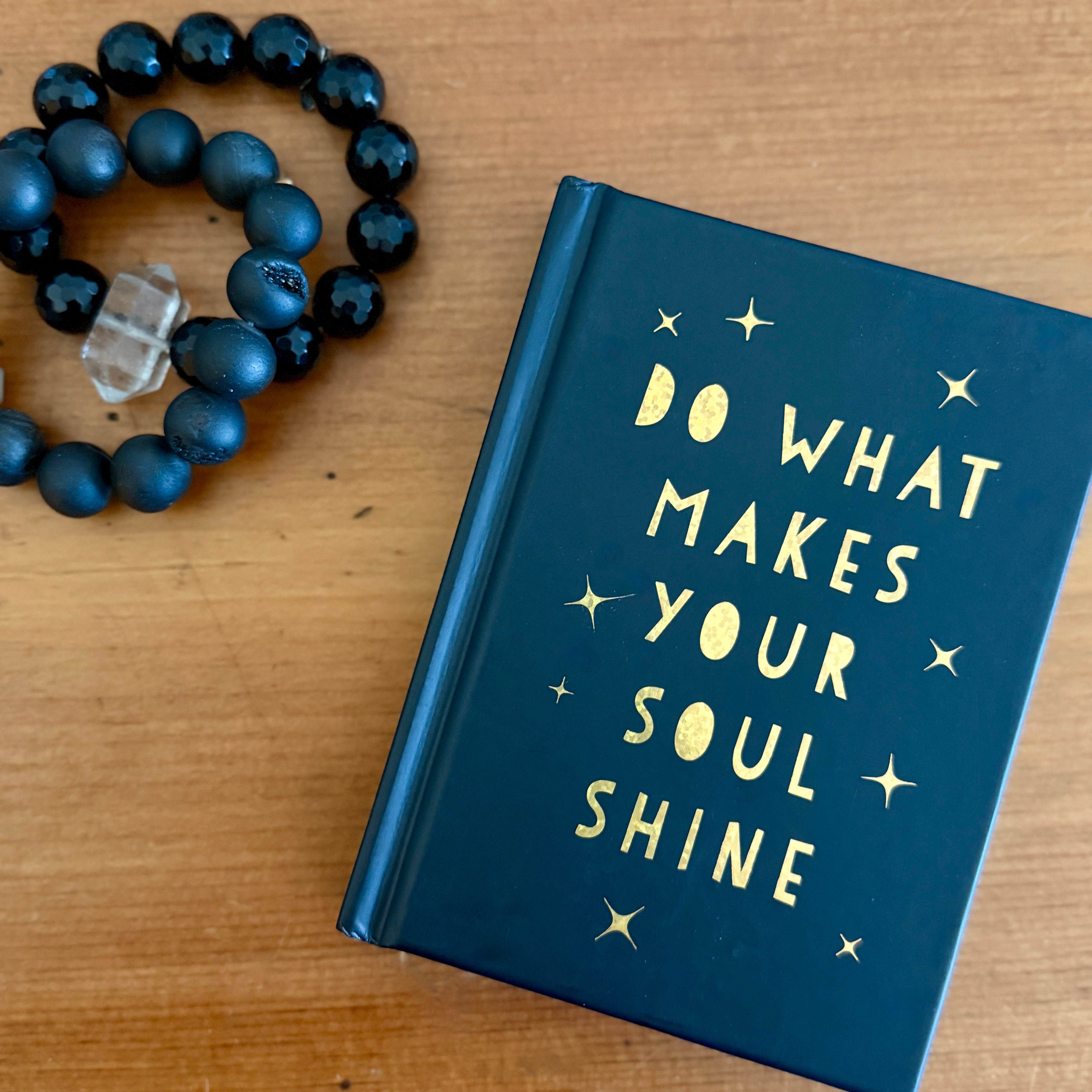 Black book with 'Do what makes your soul shine' text on a wooden surface with a black beaded bracelet.