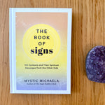Book titled 'The Book of Signs' by Mystic Michaela on a wooden surface with a purple crystal.