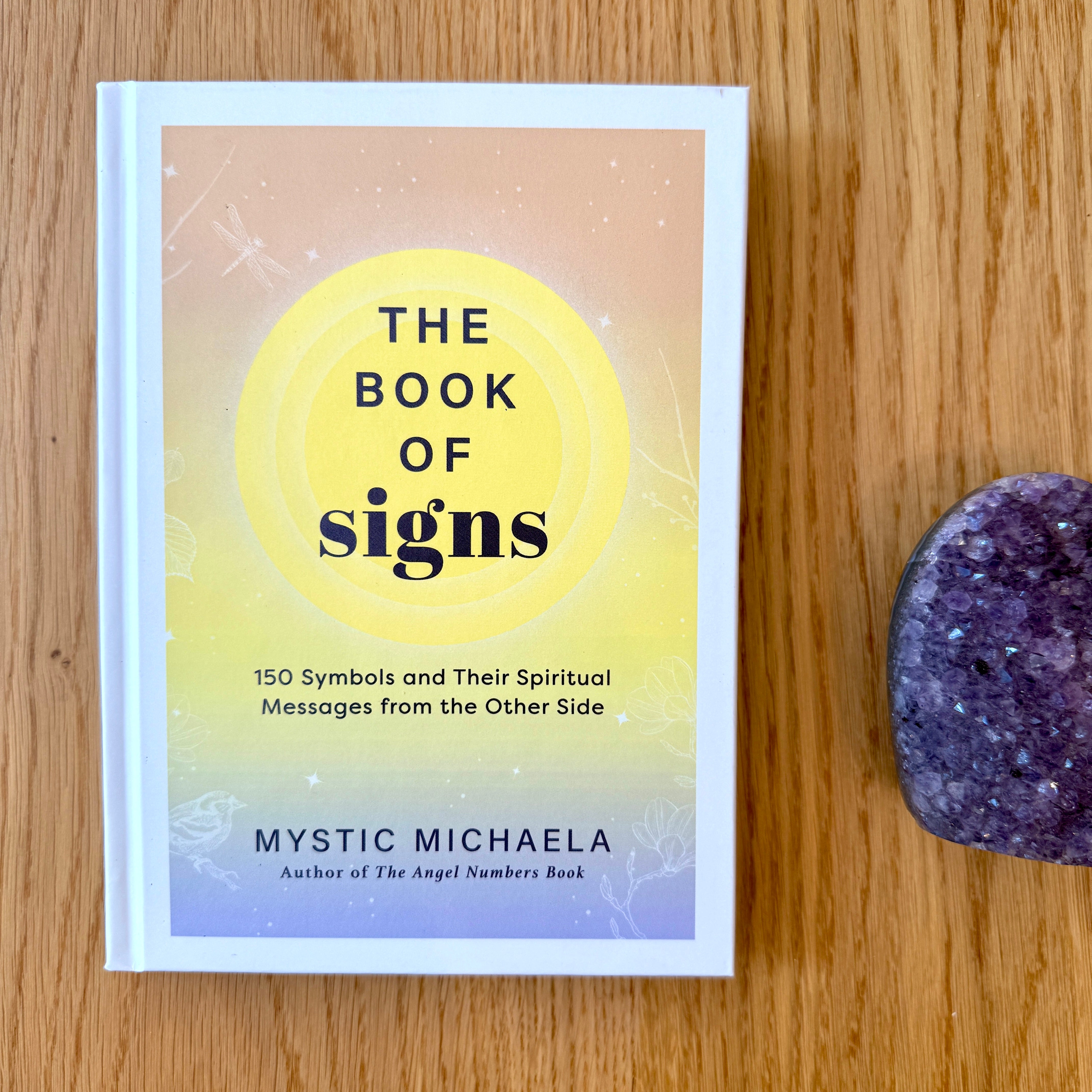 Book titled 'The Book of Signs' by Mystic Michaela on a wooden surface with a purple crystal.