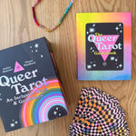 Queer Tarot cards and guidebook on a wooden surface with a colorful bracelet.