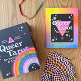 Queer Tarot cards and guidebook on a wooden surface with a colorful bracelet.