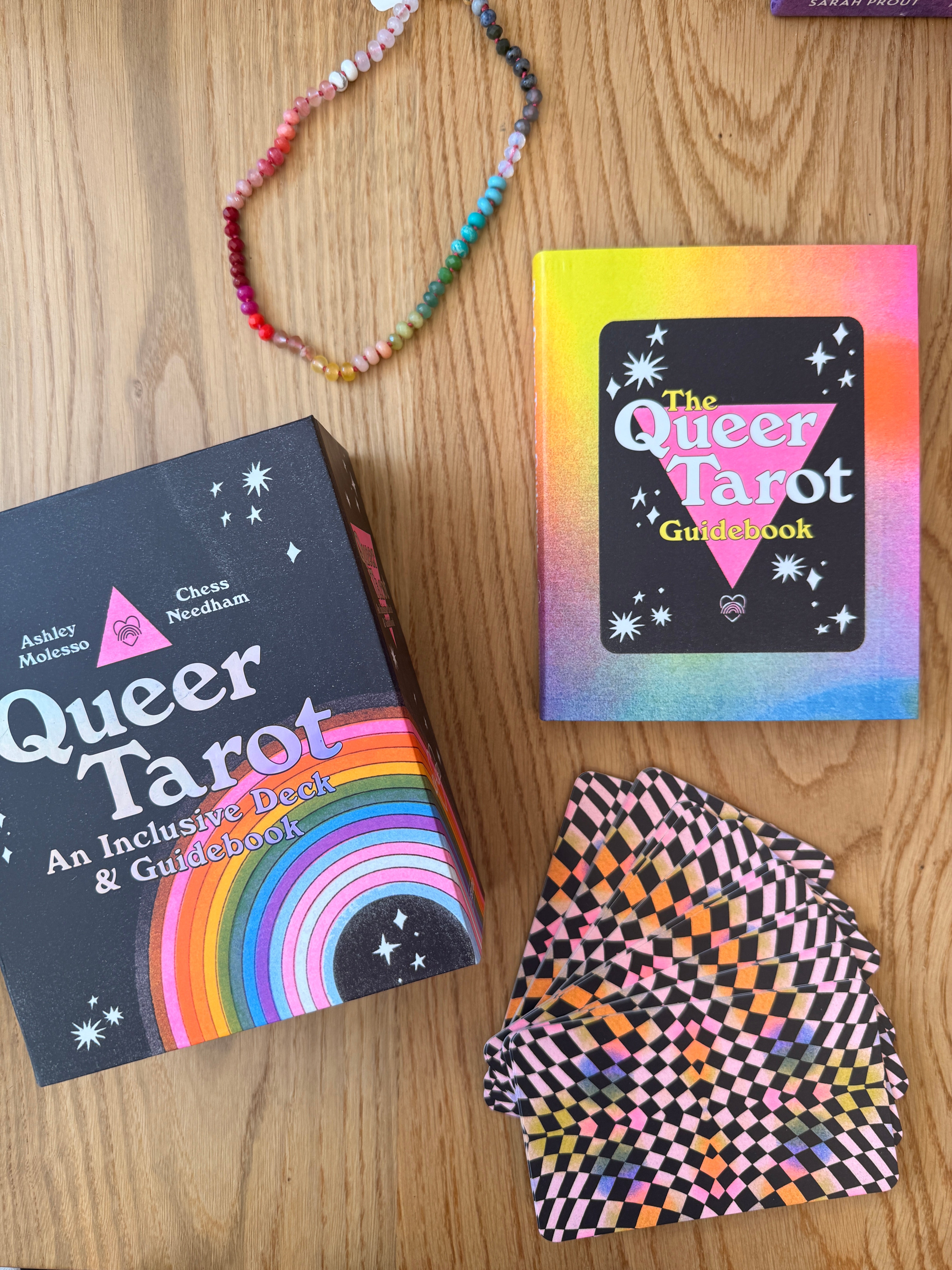 Queer Tarot cards and guidebook on a wooden surface with a colorful bracelet.