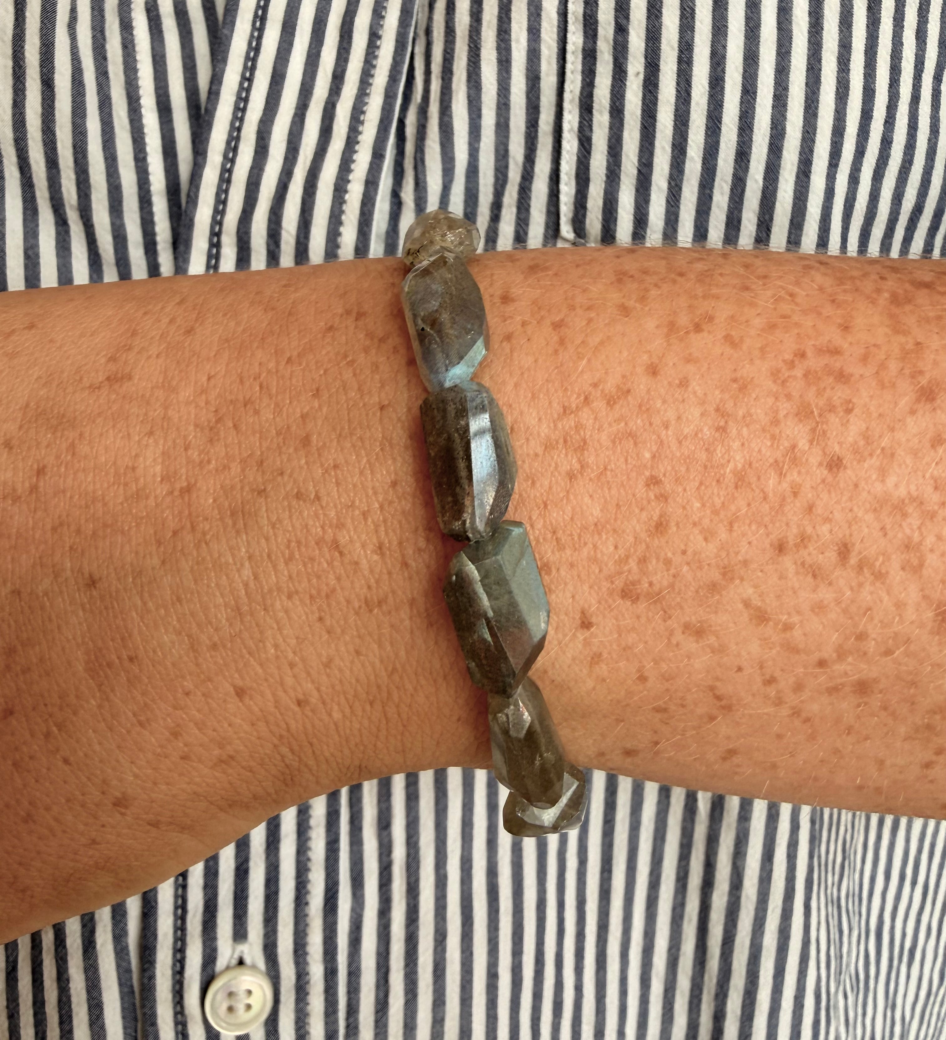 A close-up of a person's wrist wearing a beaded bracelet made of faceted dark gray stone beads, worn over a navy and white striped button-down shirt.