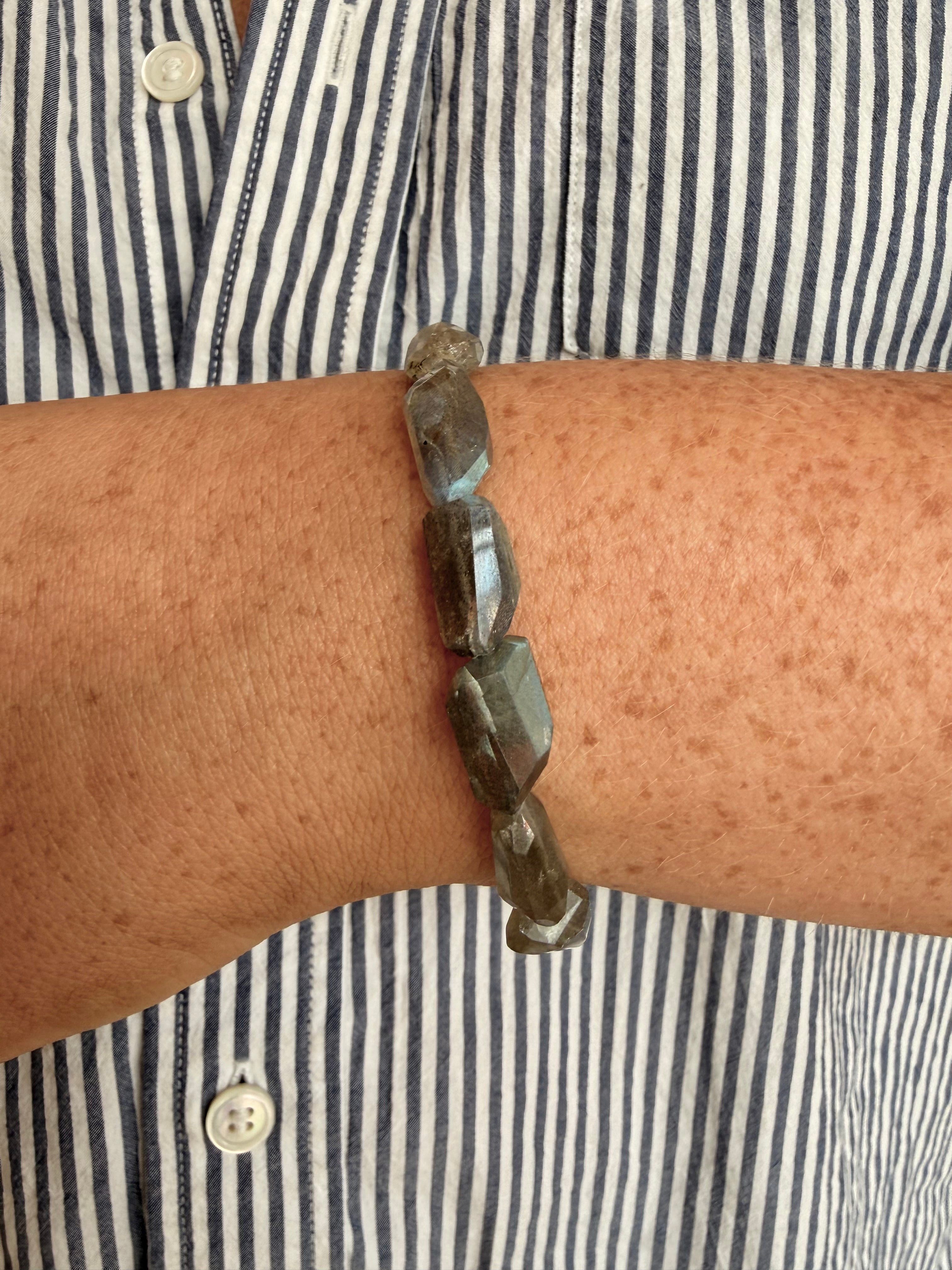 A close-up of a person's wrist wearing a beaded bracelet made of faceted dark gray stone beads, worn over a navy and white striped button-down shirt.