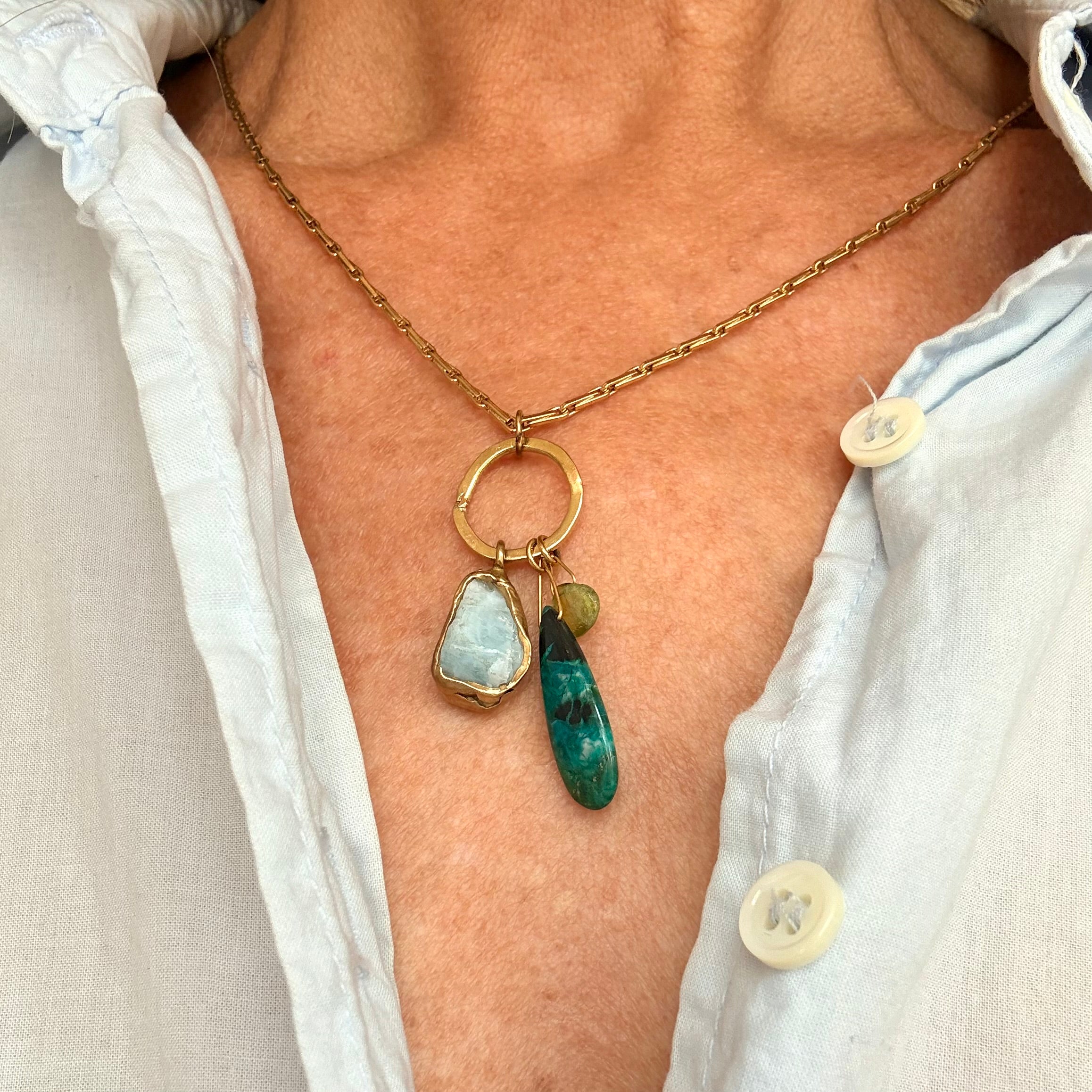 A close-up of a person wearing a gold chain necklace with a ring connector and two dangling stone pendants: one light blue stone and one green stone, styled over a light blue button-up shirt.