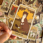 A hand holds a tarot card titled 'Strength' featuring an illustration of a brown and white cat with a flower crown, surrounded by a scattered pile of other illustrated tarot cards.