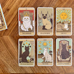 Six illustrated tarot-style cards featuring cats and a sun are arranged on a wooden tabletop. The cards include The Emperor, Six of Swords, The Sun, The High Priestess, The Star, and Queen of Wands, with part of another card visible at the upper left.