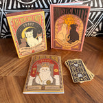 A boxed tarot deck, a tarot guide book, and several cards arranged on a wooden tabletop. The packaging and cover art feature stylized cat illustrations with ornate vintage typography reading 'Cats Rule the Earth Tarot' and 'Mystic Kitty: A Cat Guide to Meditation.