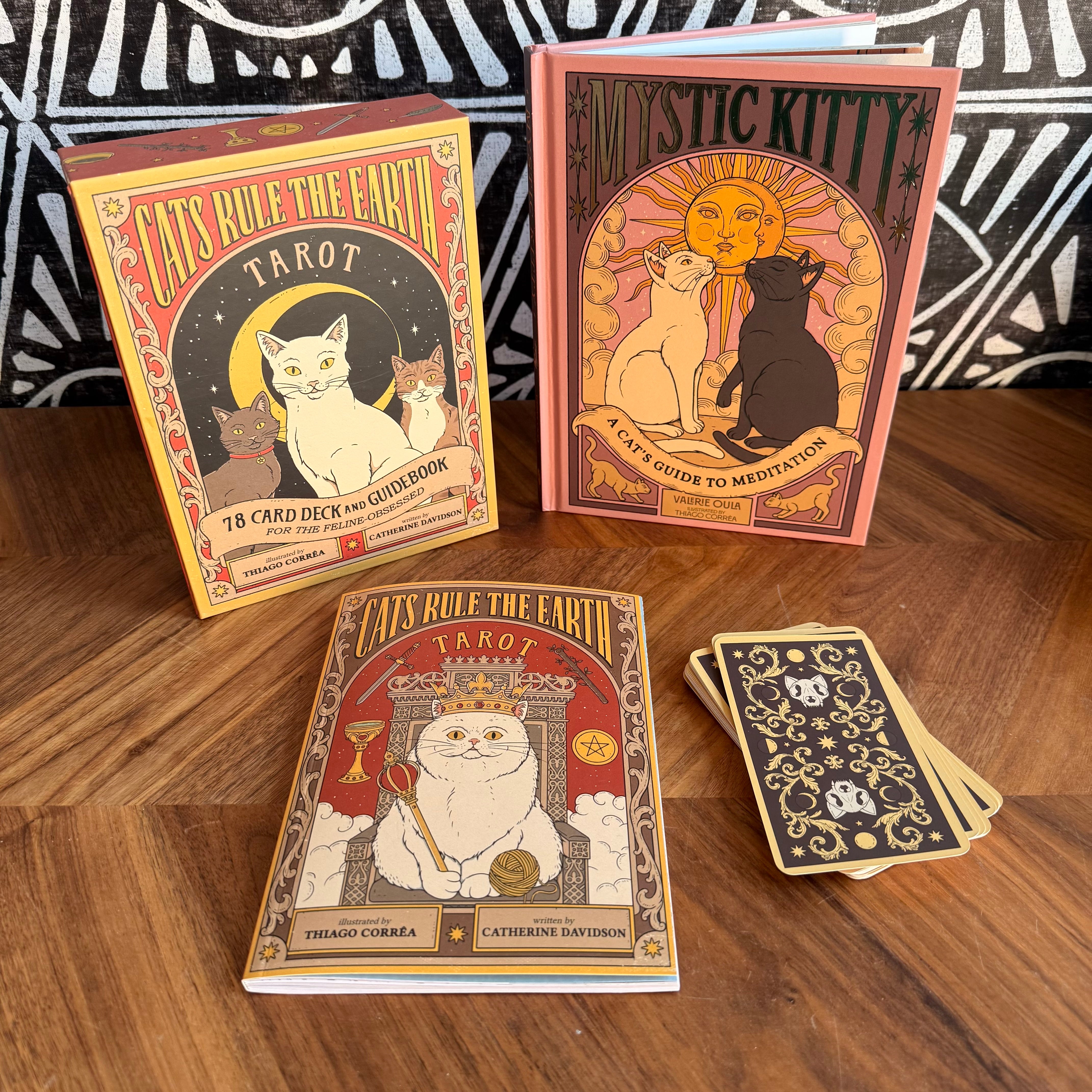 A boxed tarot deck, a tarot guide book, and several cards arranged on a wooden tabletop. The packaging and cover art feature stylized cat illustrations with ornate vintage typography reading 'Cats Rule the Earth Tarot' and 'Mystic Kitty: A Cat Guide to Meditation.