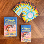 The Cosmic Crossroads Oracle deck and guidebook displayed on a wooden tabletop, with the deck fanned out to show the colorful starry sky card backs.