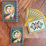 The Omen Oracle card deck and guidebook set displayed on a wooden surface, including the box, a fanned-out deck of cards, and the guidebook.