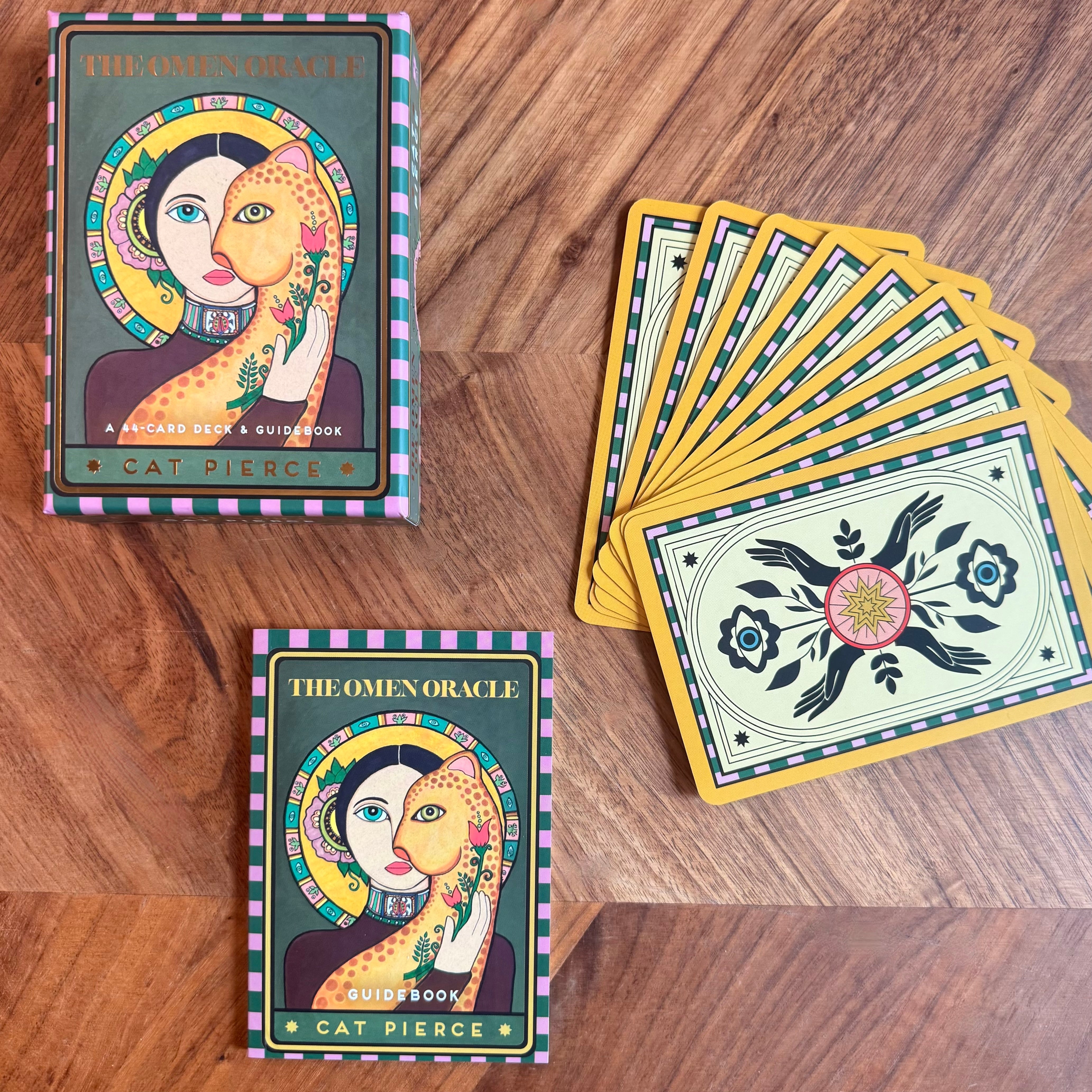 The Omen Oracle card deck and guidebook set displayed on a wooden surface, including the box, a fanned-out deck of cards, and the guidebook.