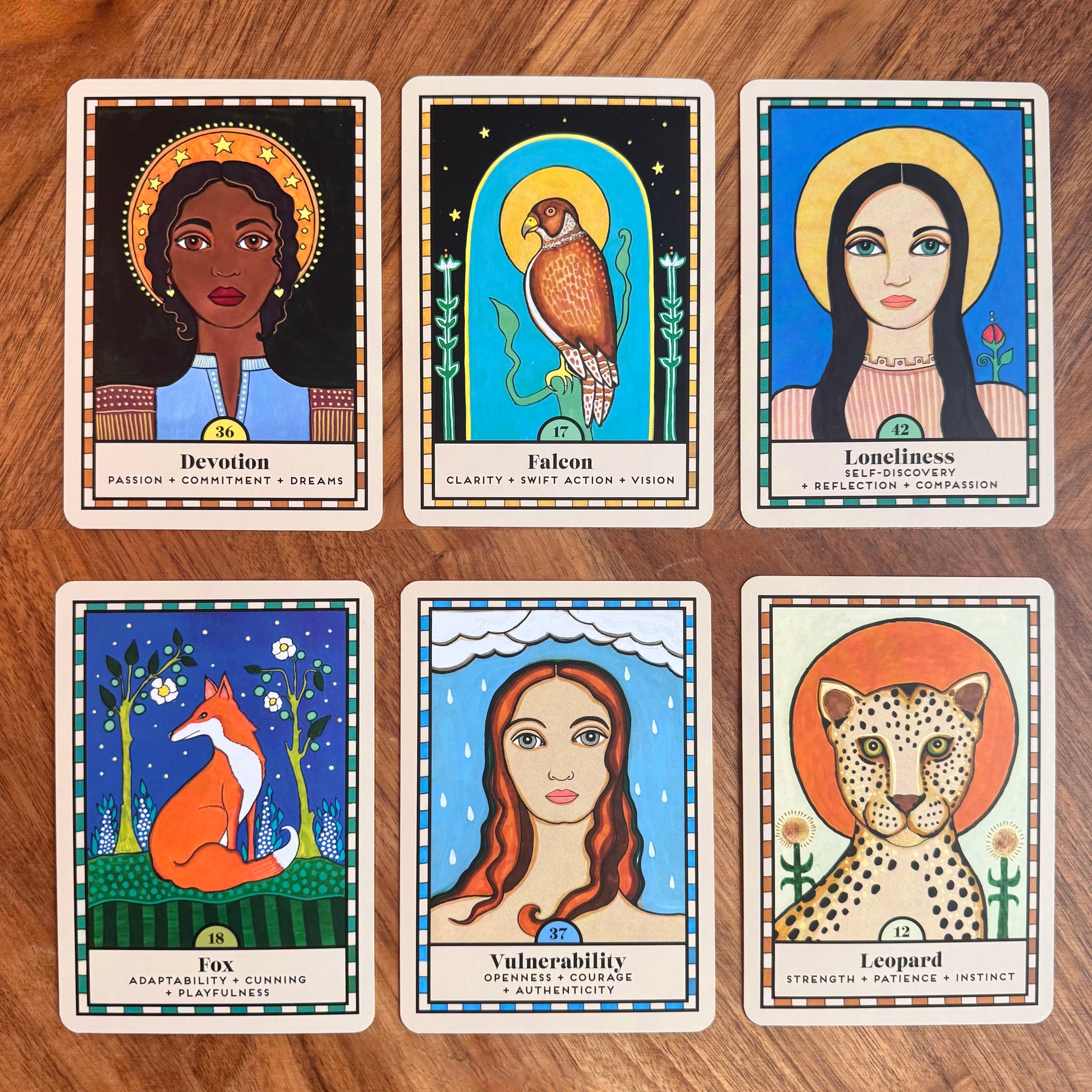 Six illustrated tarot-style cards arranged in two rows on a wooden surface. The cards feature stylized portraits and symbols with words such as Devotion, Falcon, Loneliness, Fox, Vulnerability, and Leopard.