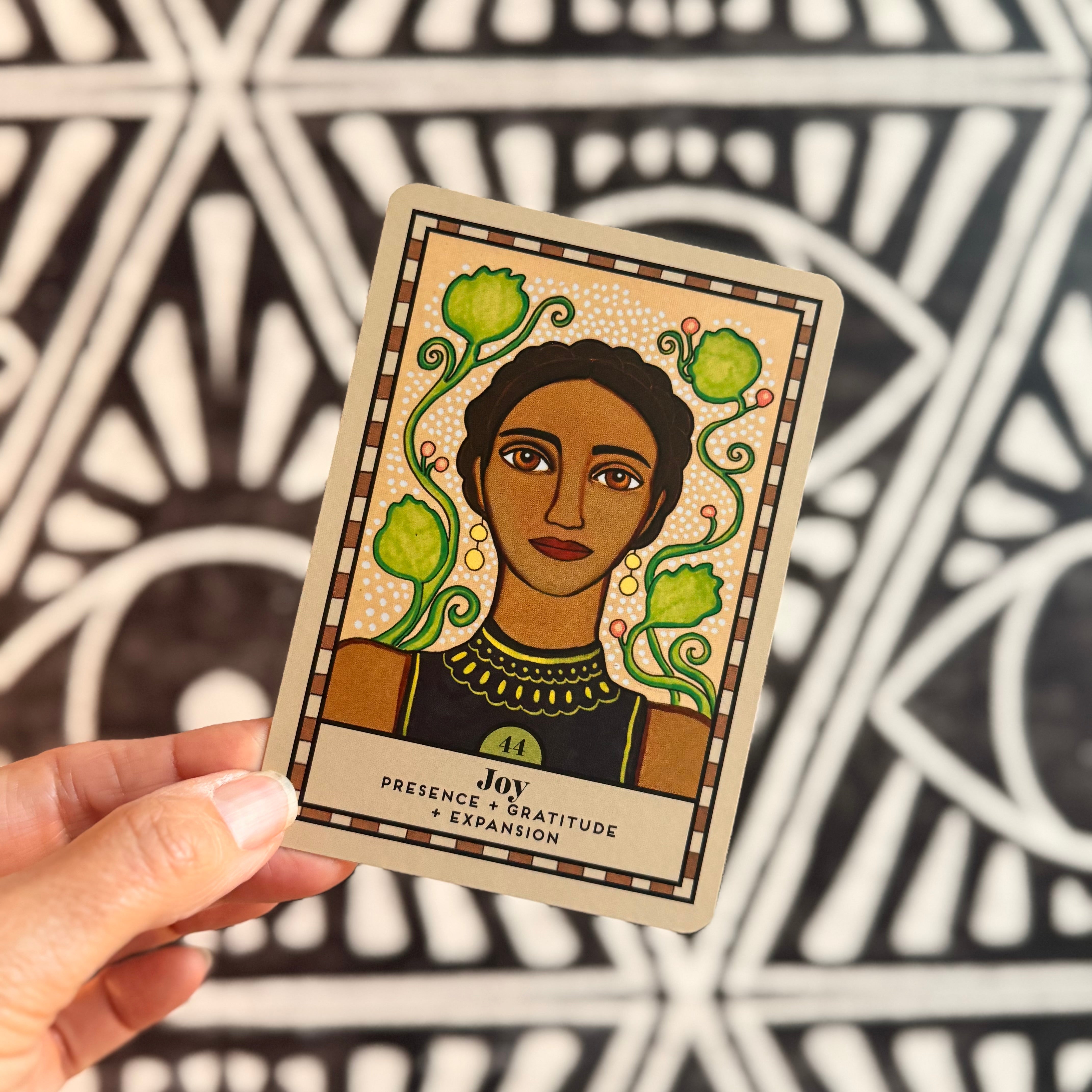 A hand holds a tarot card titled 'Joy: Presence, Gratitude & Expansion' in front of a black and white geometric patterned background. The card features an illustration of a woman with dark skin, short hair, and a black top, surrounded by green vines and flowers on a cream-colored background.
