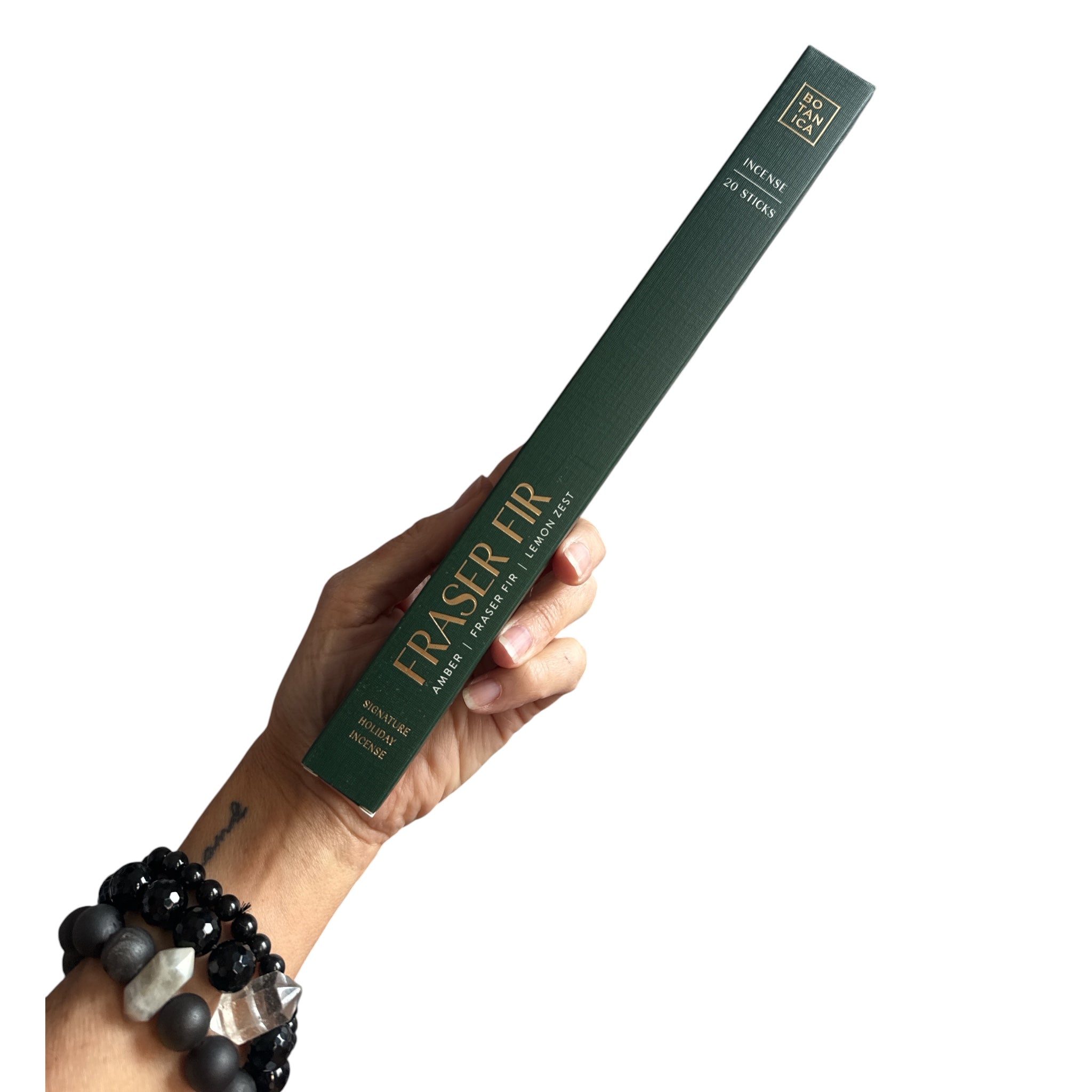 A hand holding a long, dark green box of Fraser Fir Organic Lip Balm against a solid white background. The box features gold lettering and branding, and the hand is adorned with black and gray beaded bracelets.