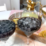 Two stemmed coupe glasses filled with crushed ice and a dark purple-black liquid, set on a table with a gold-toned decanter and tray in the background.