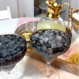 Two stemmed coupe glasses filled with crushed ice and a dark purple-black liquid, set on a table with a gold-toned decanter and tray in the background.
