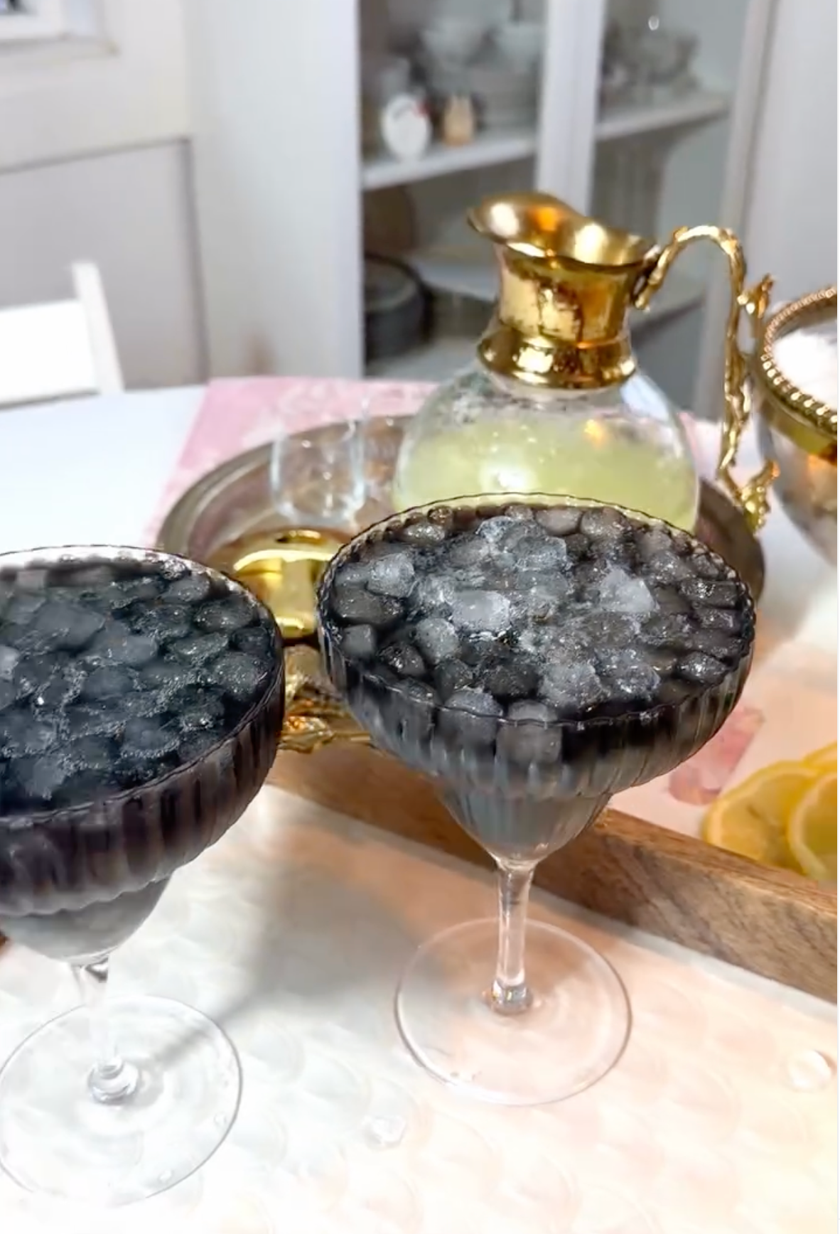 Two stemmed coupe glasses filled with crushed ice and a dark purple-black liquid, set on a table with a gold-toned decanter and tray in the background.