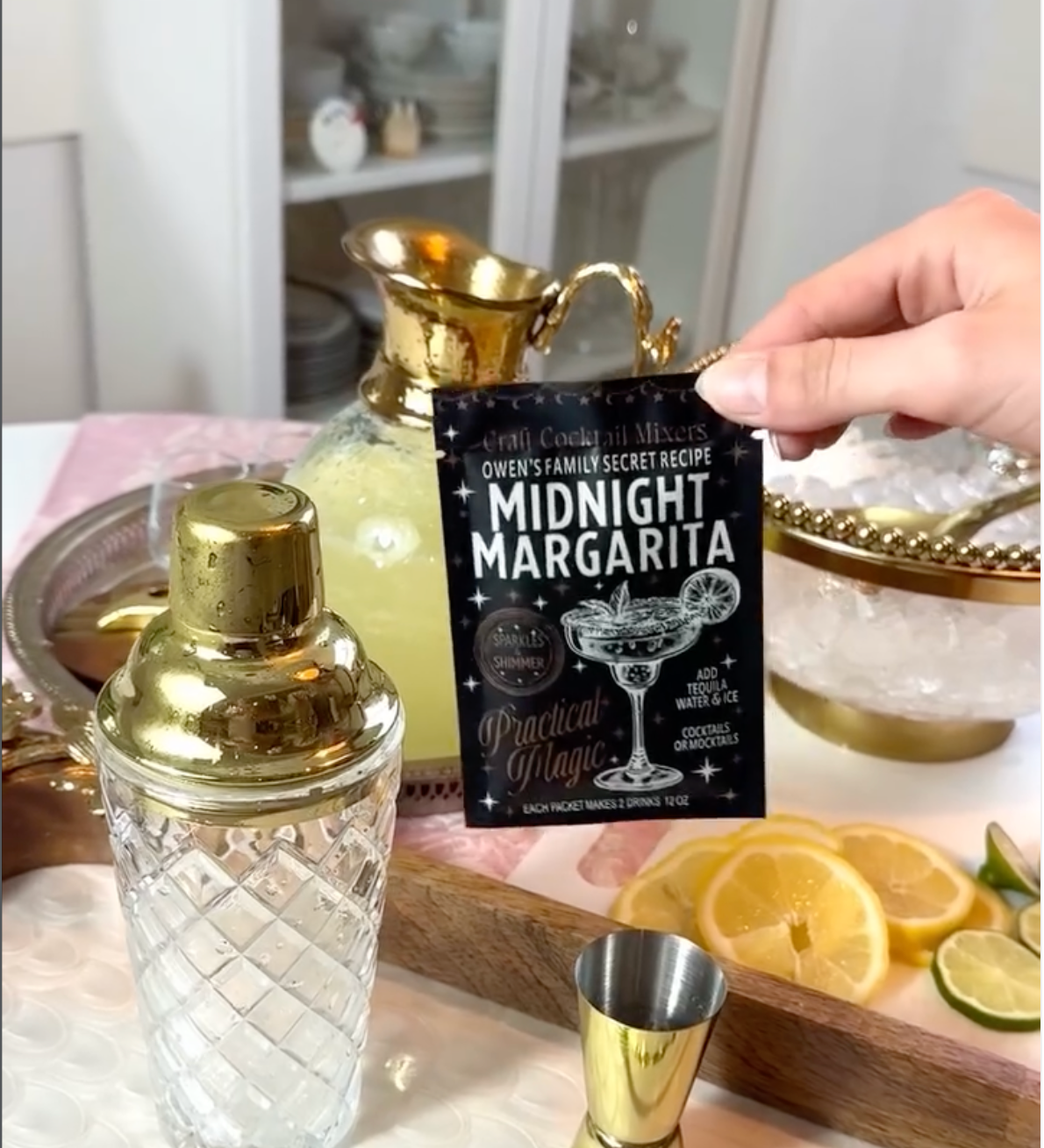 A hand holds a black card reading 'Queen's Family Secret Recipe: Midnight Margarita' in front of a bar-style setup with a gold shaker, gold jigger, ice bowl, and lemon slices.
