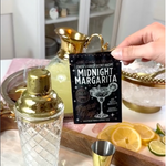 A hand holds a black card reading 'Queen's Family Secret Recipe: Midnight Margarita' in front of a bar-style setup with a gold shaker, gold jigger, ice bowl, and lemon slices.