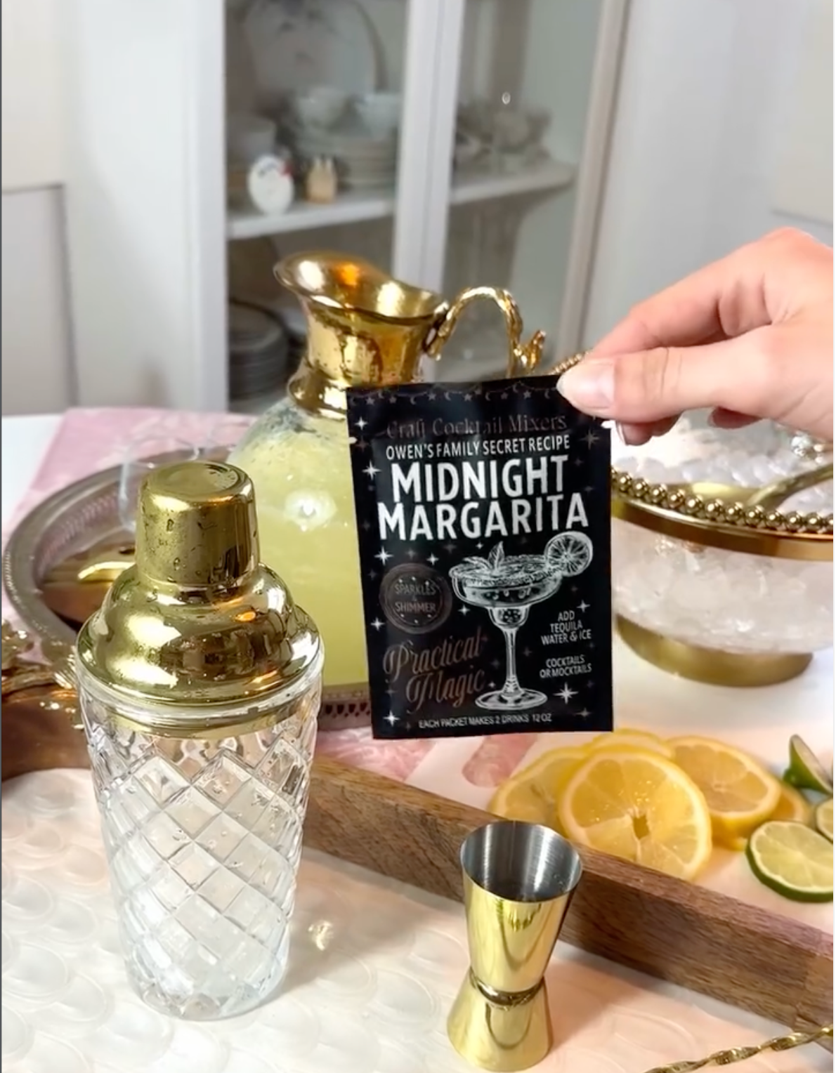 A hand holds a black card reading 'Queen's Family Secret Recipe: Midnight Margarita' in front of a bar-style setup with a gold shaker, gold jigger, ice bowl, and lemon slices.