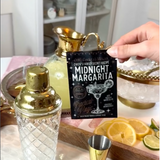 A hand holds a black card reading 'Queen's Family Secret Recipe: Midnight Margarita' in front of a bar-style setup with a gold shaker, gold jigger, ice bowl, and lemon slices.