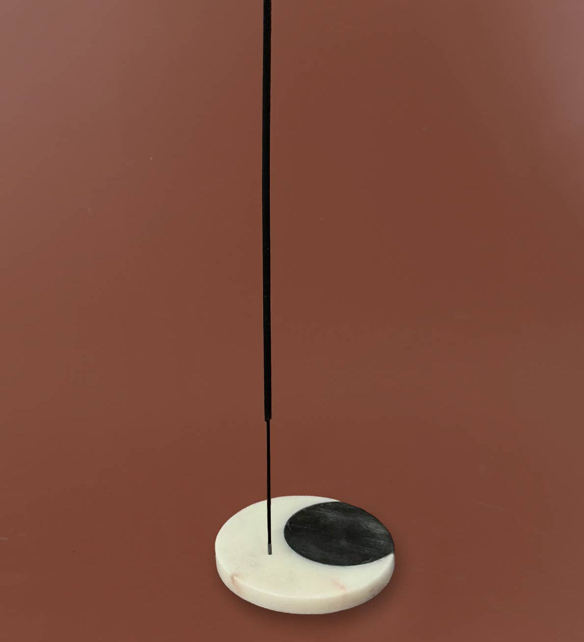 A minimalist incense holder made of a white marble base with a black semicircle on one side, holding a single vertical black incense stick, set against a solid terracotta background.