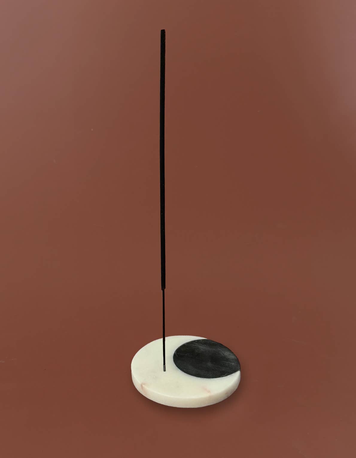 A minimalist incense holder made of a white marble base with a black semicircle on one side, holding a single vertical black incense stick, set against a solid terracotta background.