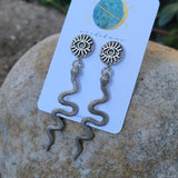 Pair of silver snake-shaped earrings on a card with 'veridilum' branding, placed on a stone surface.