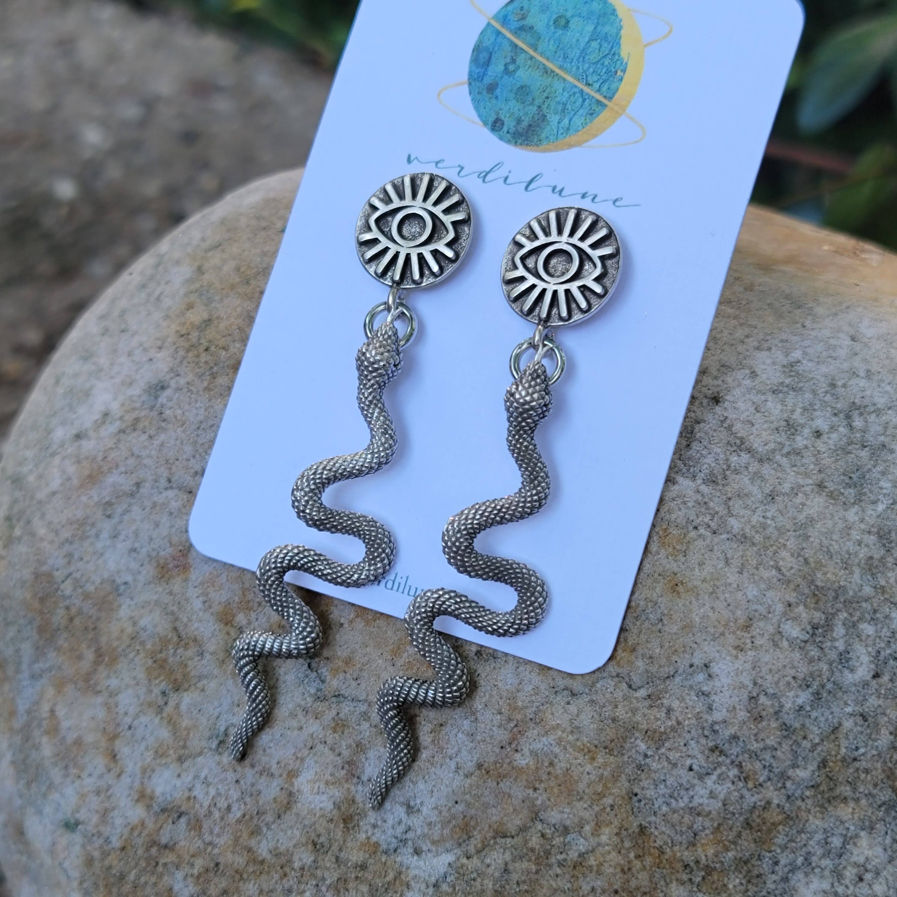 Pair of silver snake-shaped earrings on a card with 'veridilum' branding, placed on a stone surface.
