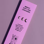 A tall, narrow lavender-pink product box for Brass Earth, featuring black text and illustrations including a rabbit and moon phases, photographed on a matching lavender background.