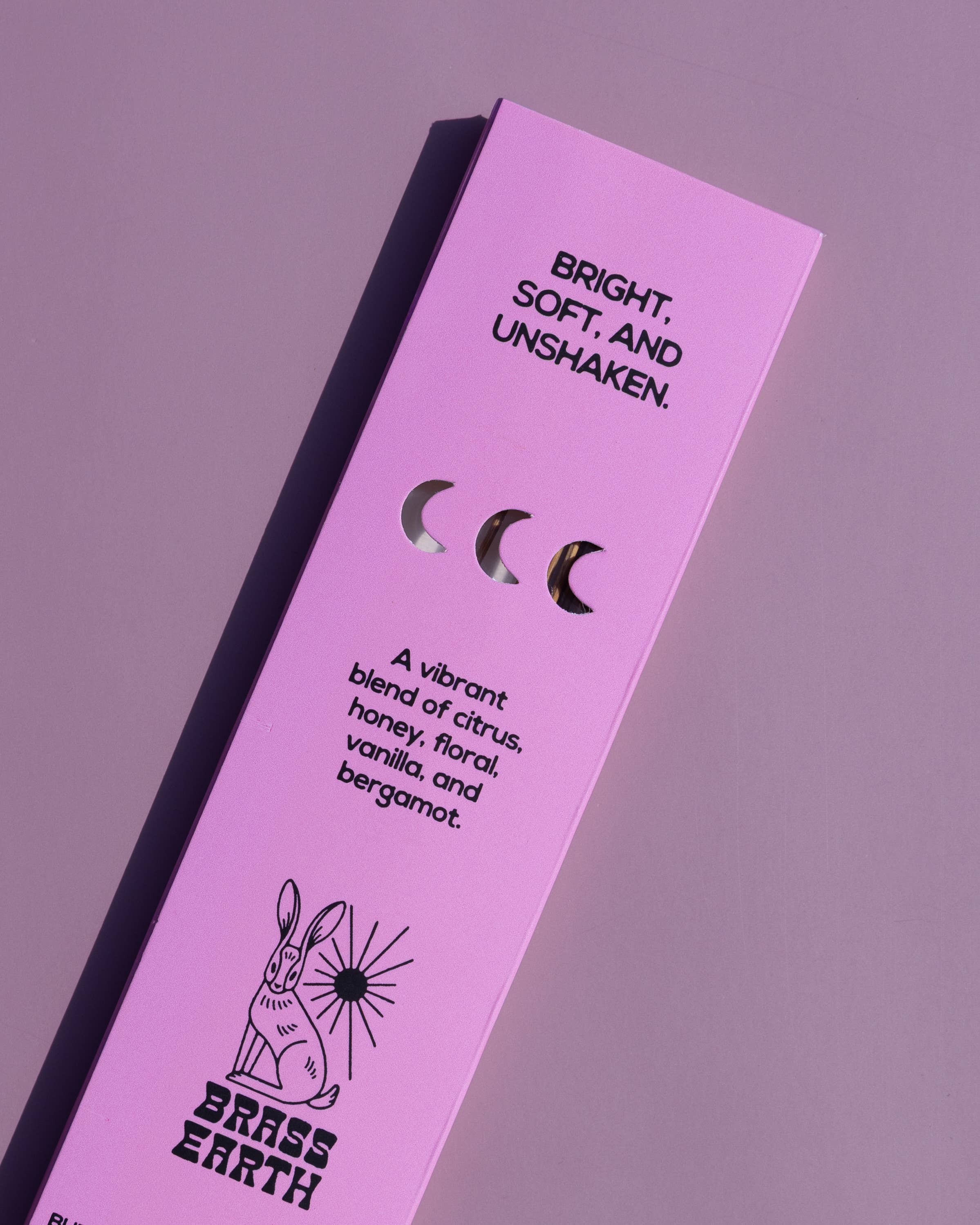 A tall, narrow lavender-pink product box for Brass Earth, featuring black text and illustrations including a rabbit and moon phases, photographed on a matching lavender background.