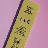 A rectangular brass candle tin by Brass Earth, shown diagonally on a solid pink background. The tin is a muted yellow-green color with black text and illustrations, including a rabbit and moon phases.