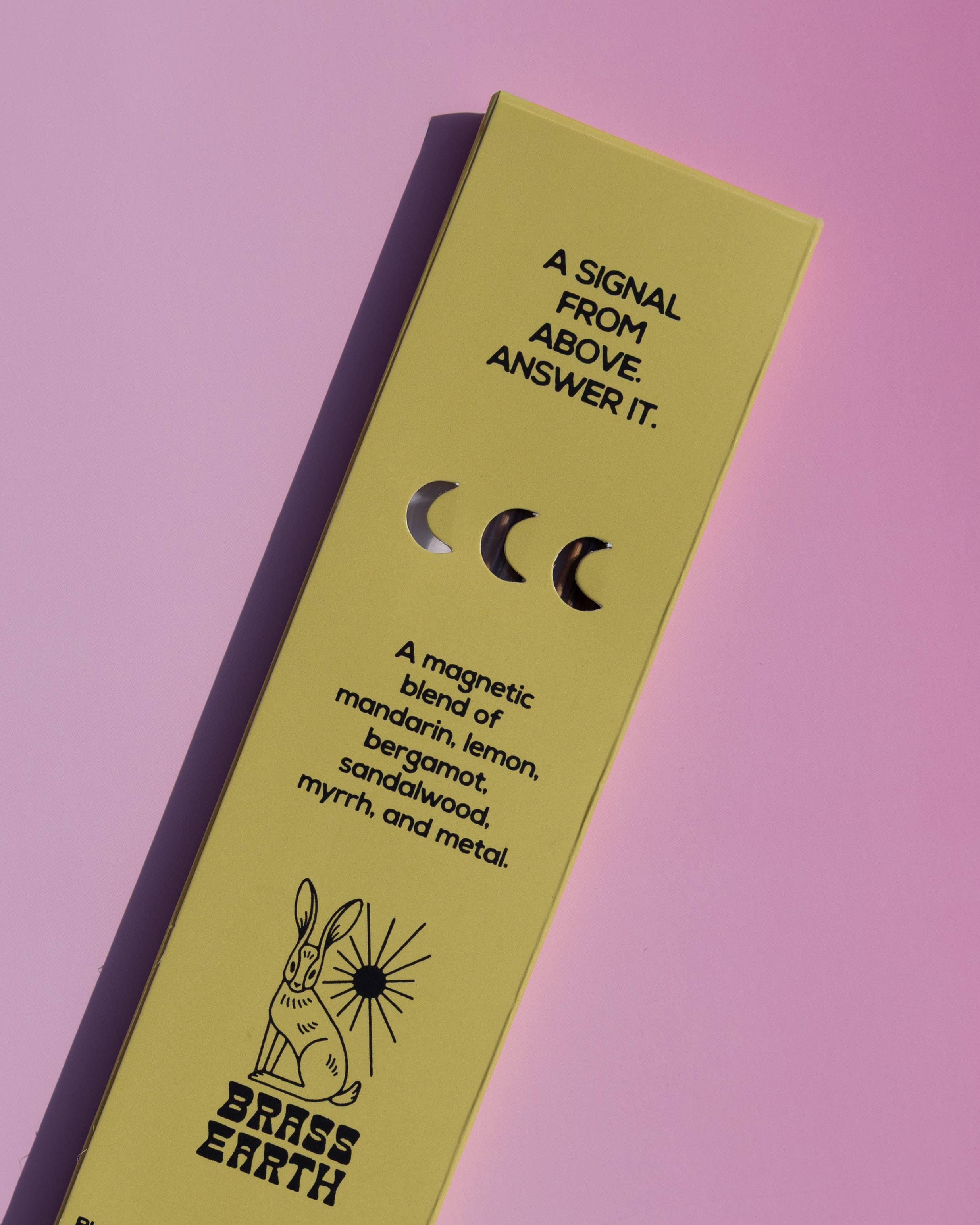 A rectangular brass candle tin by Brass Earth, shown diagonally on a solid pink background. The tin is a muted yellow-green color with black text and illustrations, including a rabbit and moon phases.