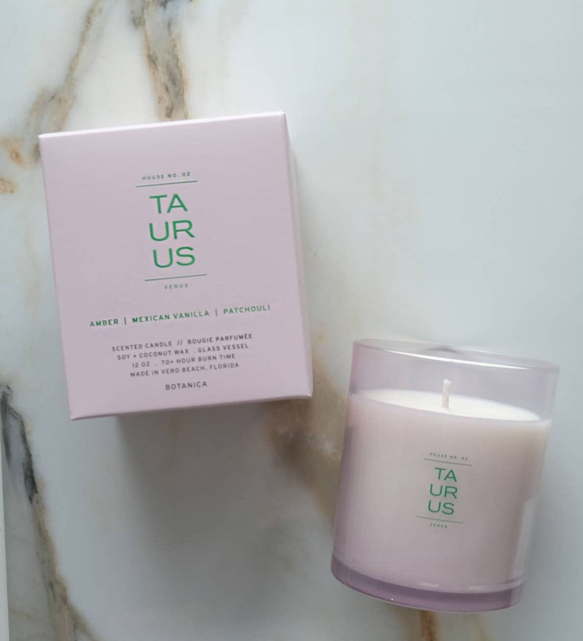 A Botanica 'Taurus' scented candle in a clear glass jar next to its light pink packaging box, displayed on a white marble surface.