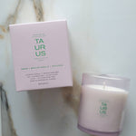 A Botanica 'Taurus' scented candle in a clear glass jar next to its light pink packaging box, displayed on a white marble surface.