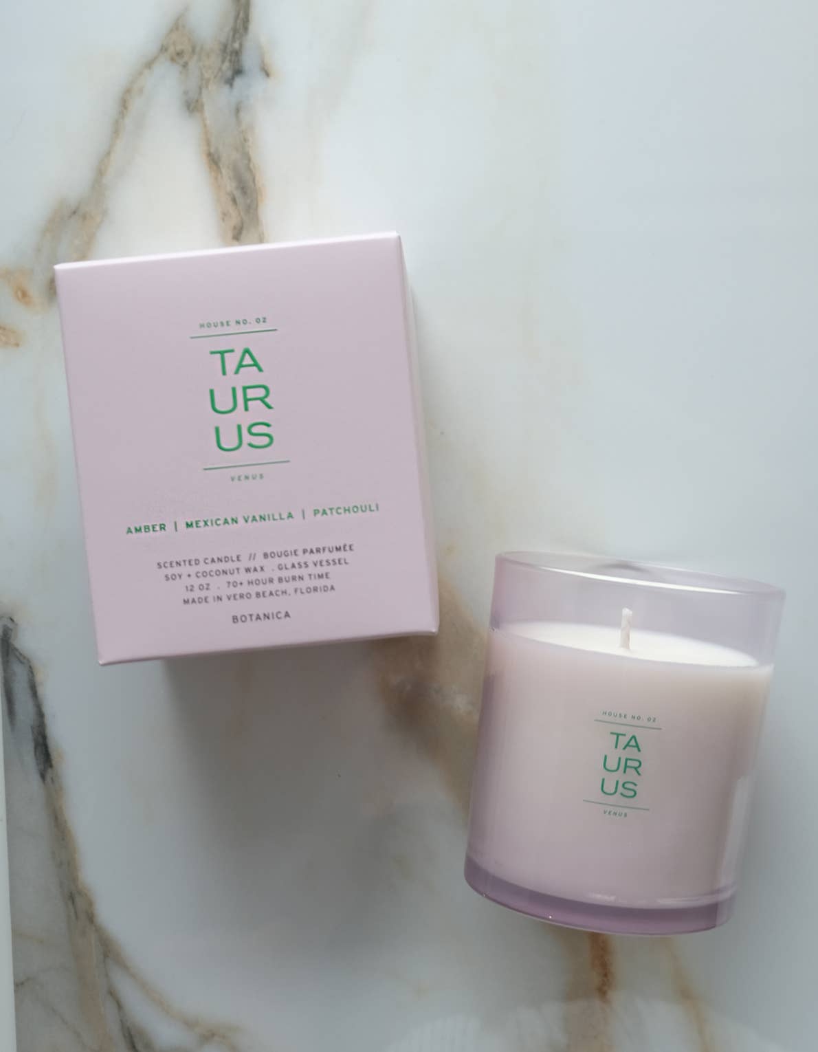 A Botanica 'Taurus' scented candle in a clear glass jar next to its light pink packaging box, displayed on a white marble surface.