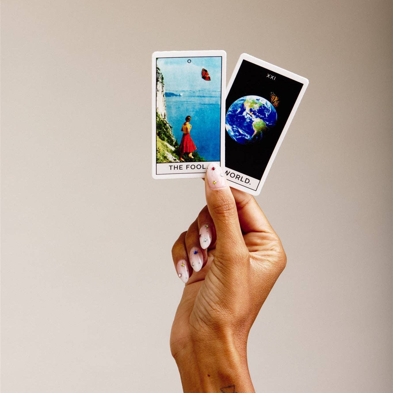A hand with light pink manicured nails holds two tarot cards against a plain beige background. The card on the left is 'The Fool' showing a figure by the sea, and the card on the right is 'World' showing a globe.