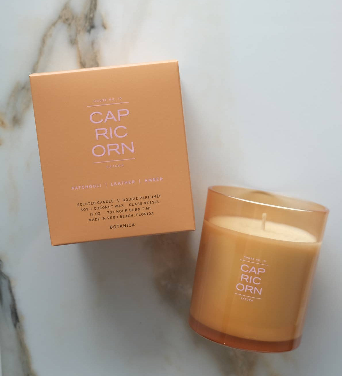 A lit scented candle in a frosted amber glass jar sits next to its peach-colored cardboard box on a light marble surface. The product label reads CAP RIC ORN.