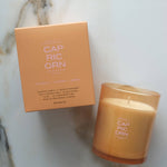 A lit scented candle in a frosted amber glass jar sits next to its peach-colored cardboard box on a light marble surface. The product label reads CAP RIC ORN.