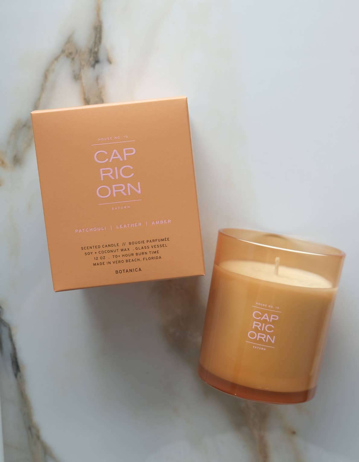 A lit scented candle in a frosted amber glass jar sits next to its peach-colored cardboard box on a light marble surface. The product label reads CAP RIC ORN.
